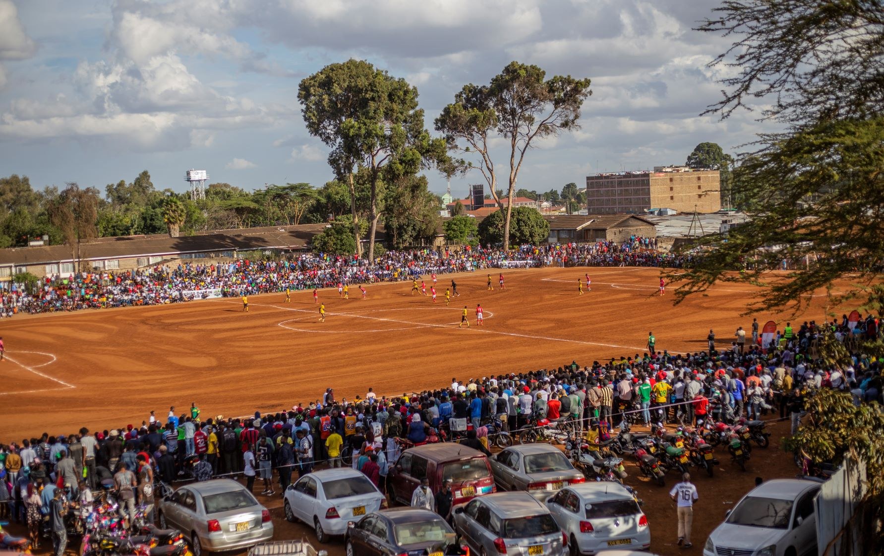 A large community crowd watches a grassroots football match on a local dirt pitch.